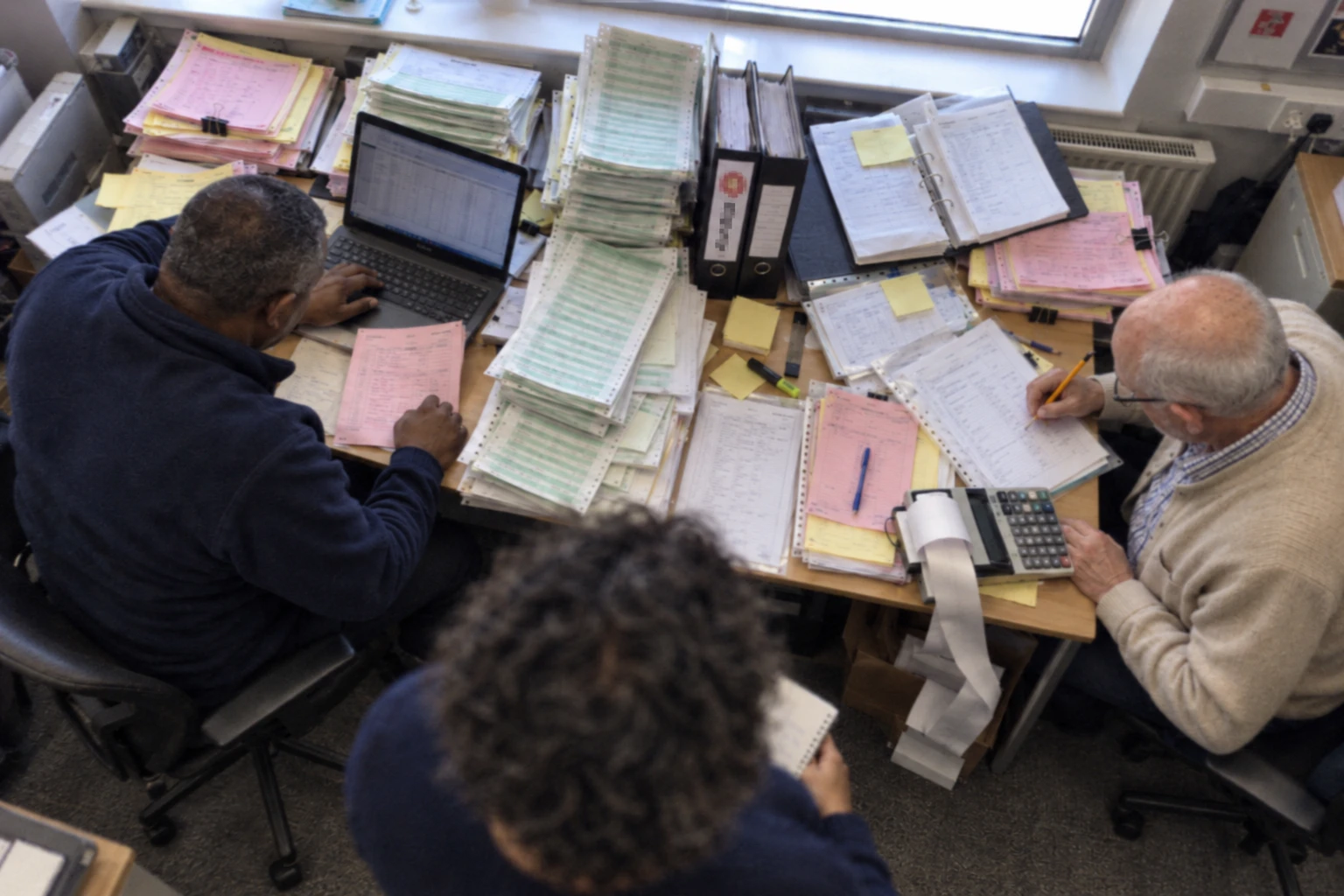 Two members of staff at an office desk stacked with printed batch sheets, lever arch files and a printing calculator, with the project manager standing behind them taking notes.