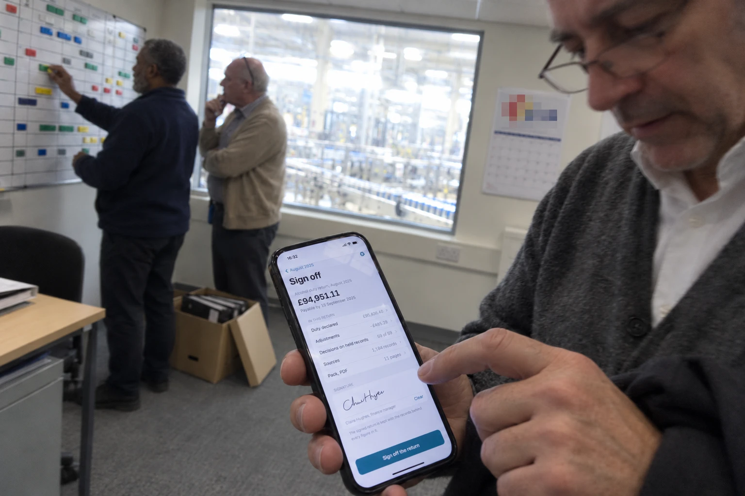 A finance manager signing off the month's duty return on his phone in an office that overlooks the production hall, with two colleagues at a planning board behind him.