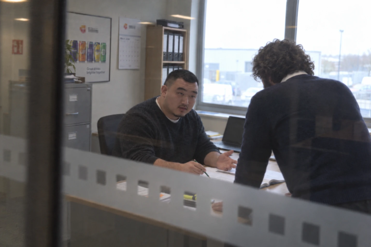 The engineer at a desk seen through a glass office partition, explaining a printed pack to the project manager, who leans over the desk with his back to the camera.